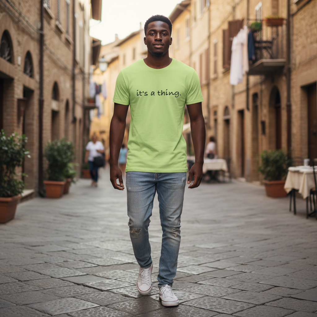 Man walking confidently down a charming cobblestone street wearing a lime green “it’s a thing.” T-shirt and jeans, showcasing a relaxed urban lifestyle and minimalist positive design.