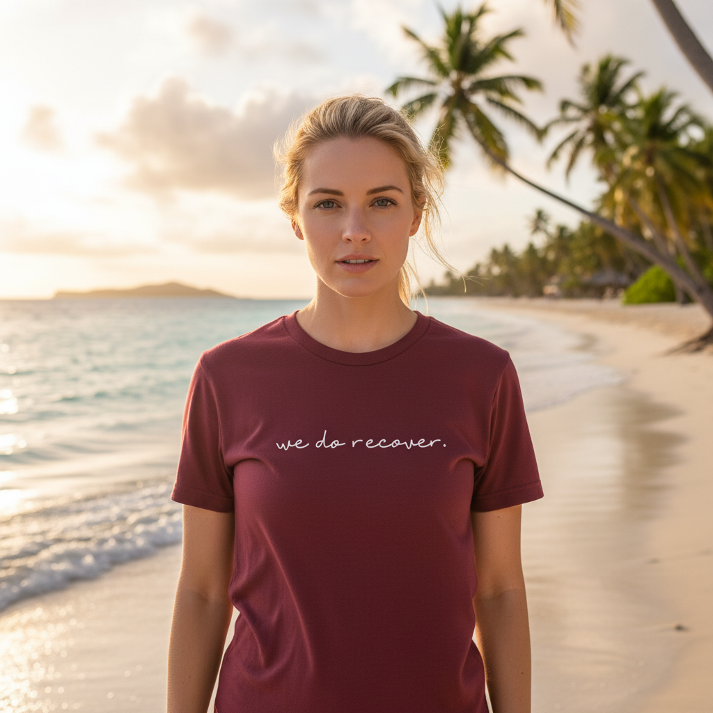 Woman standing on a tropical beach at sunset wearing a maroon T-shirt with the phrase “we do recover.” printed on the front in white text, symbolizing hope, healing, and resilience