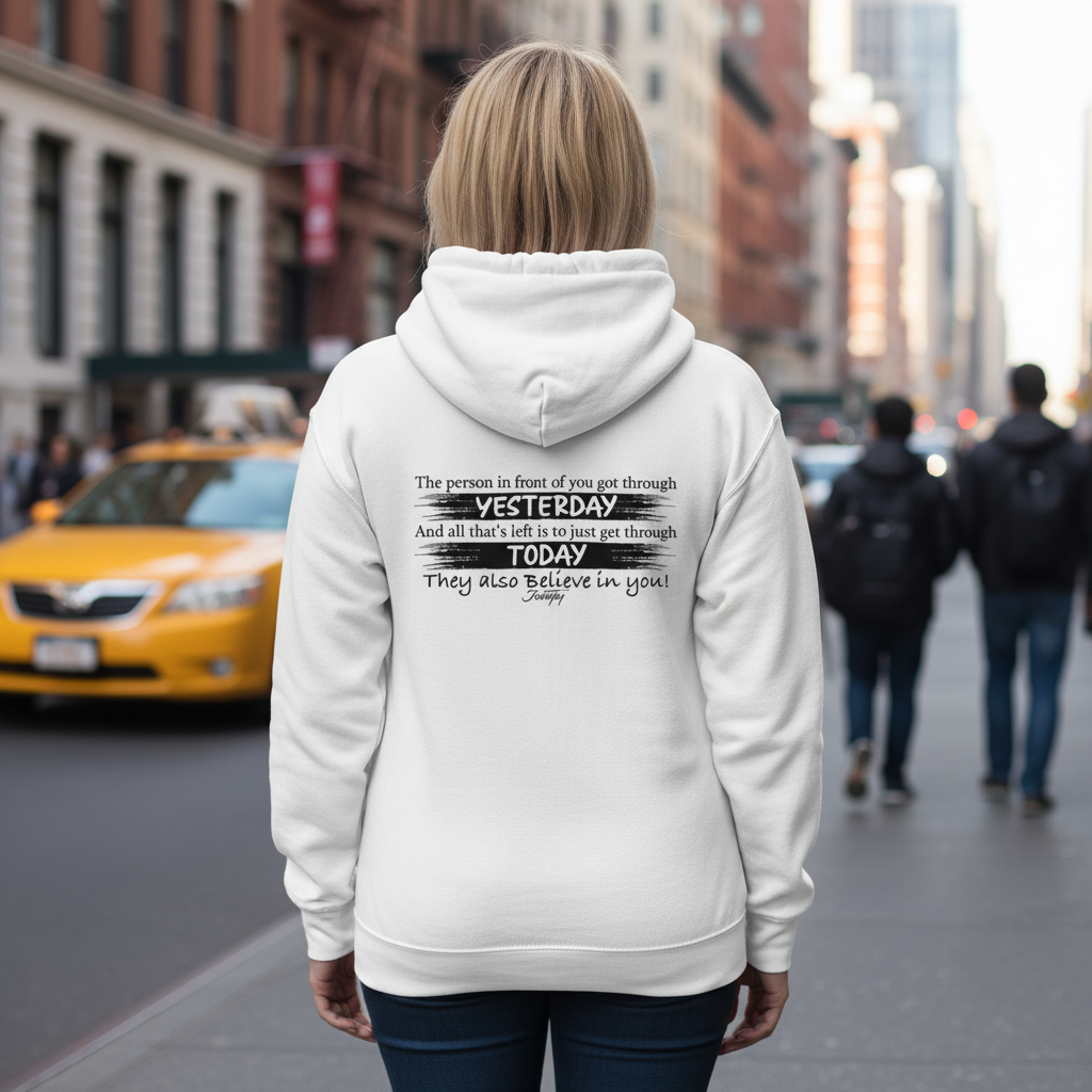 Woman walking in the city wearing a white hoodie with motivational text that reads “The person in front of you got through yesterday... They also believe in you!” symbolizing encouragement, resilience, and daily inspiration